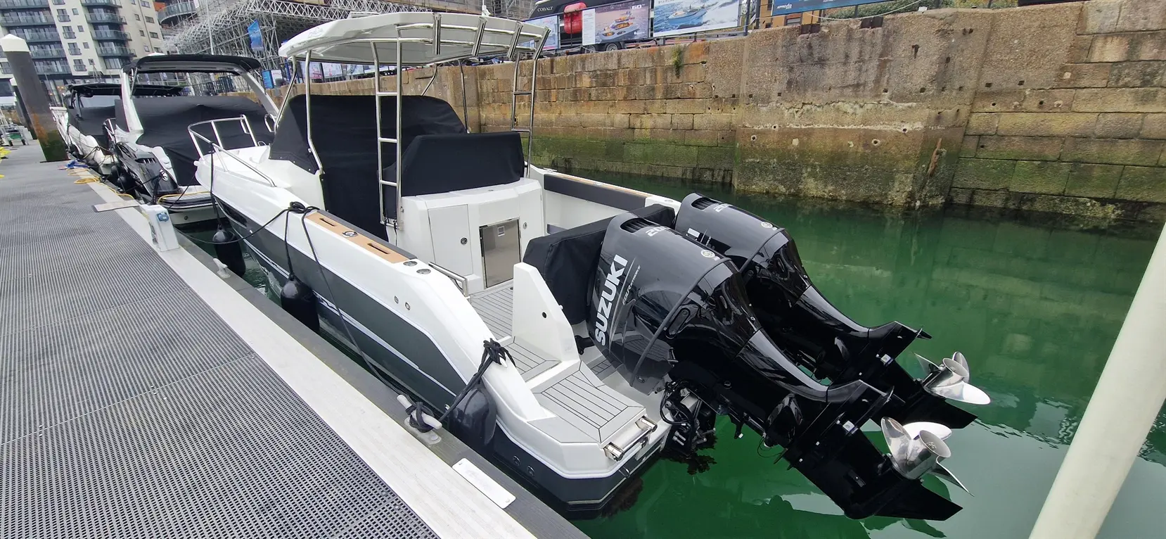 A white boat with two black Suzuki outboard motors docked beside other boats in a calm marina, surrounded by green water.